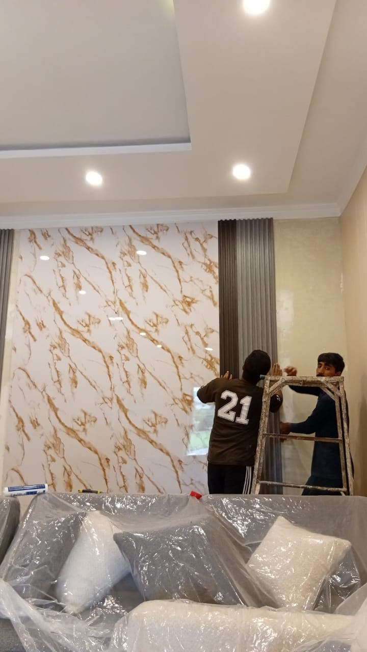 Two workers fitting a gold-veined marble-effect sheet to a lounge wall, sofas wrapped in plastic