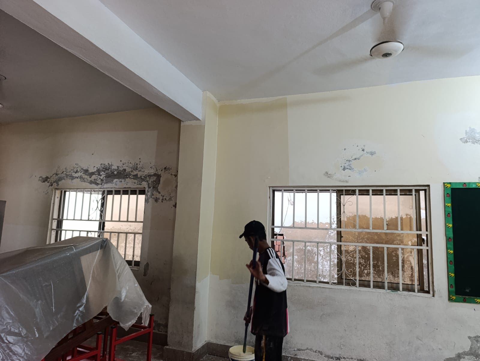 A painter rolling paint below damaged plaster, desks covered with plastic sheeting