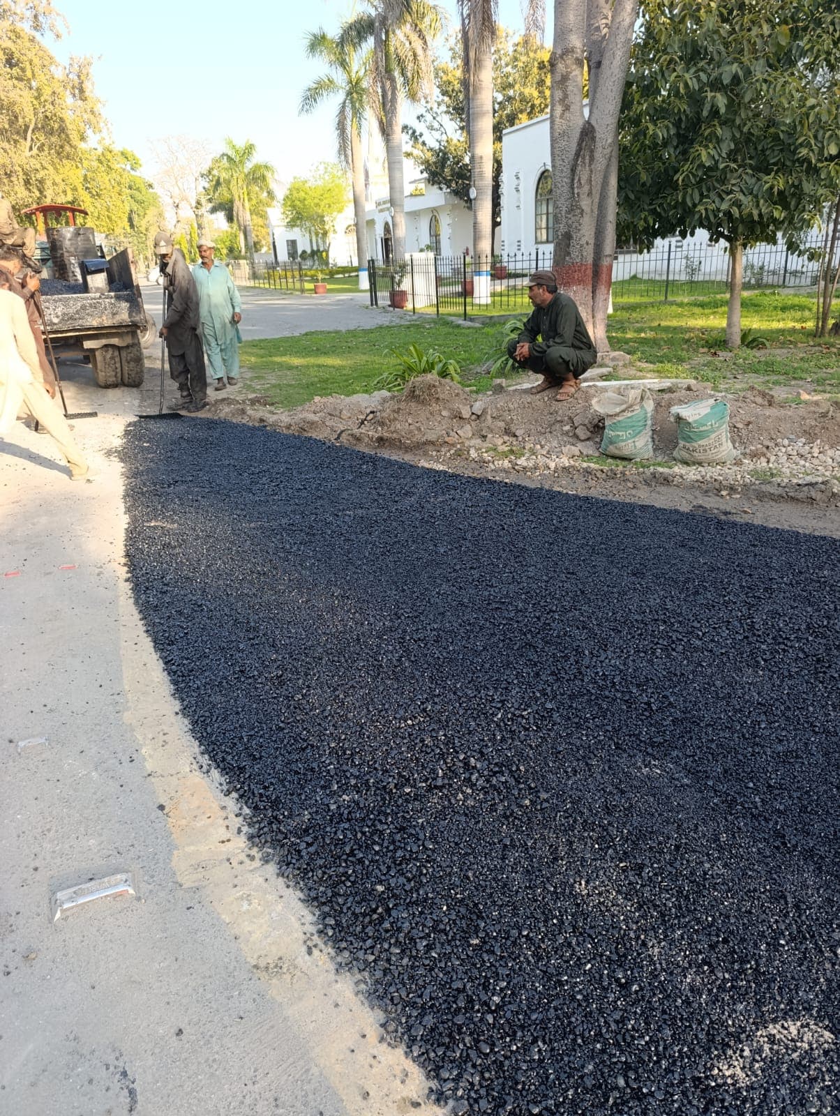 Loose asphalt spread along a road edge next to a lawn, with workers and a tractor behind