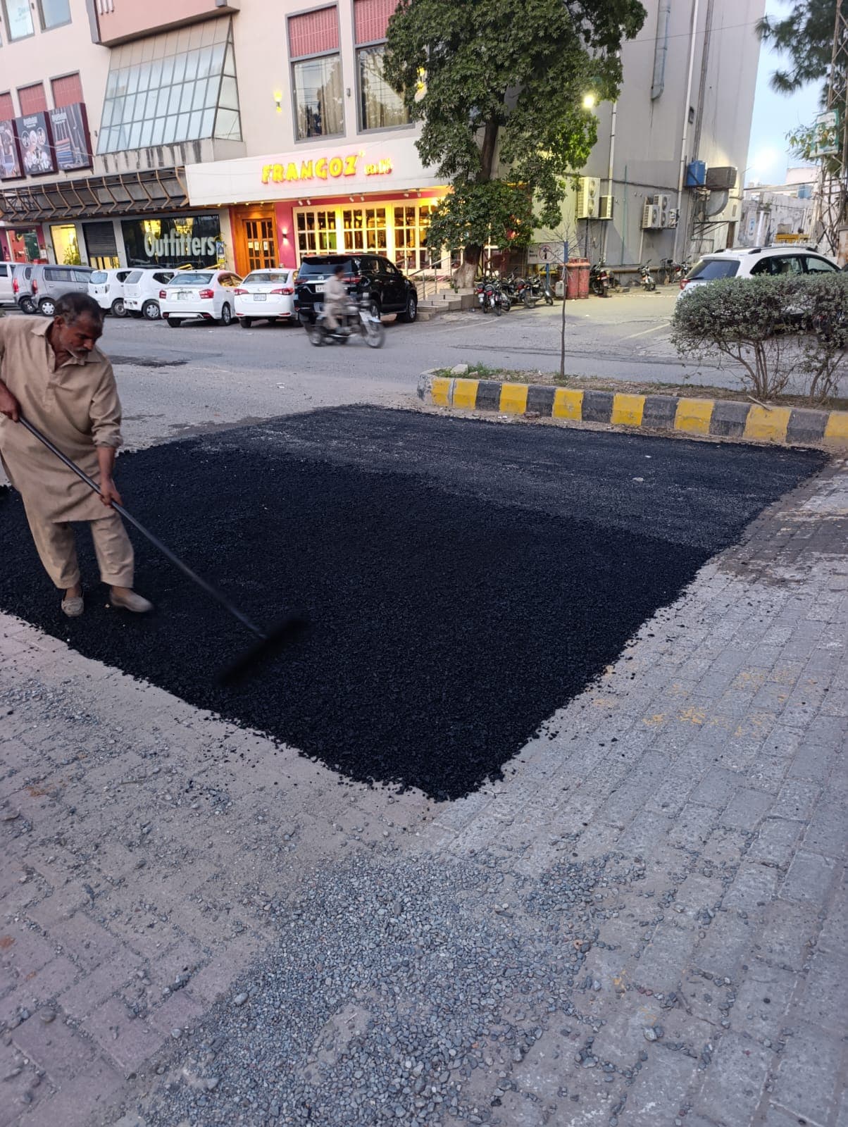 A worker raking asphalt into a patch at a market corner beside paving tiles