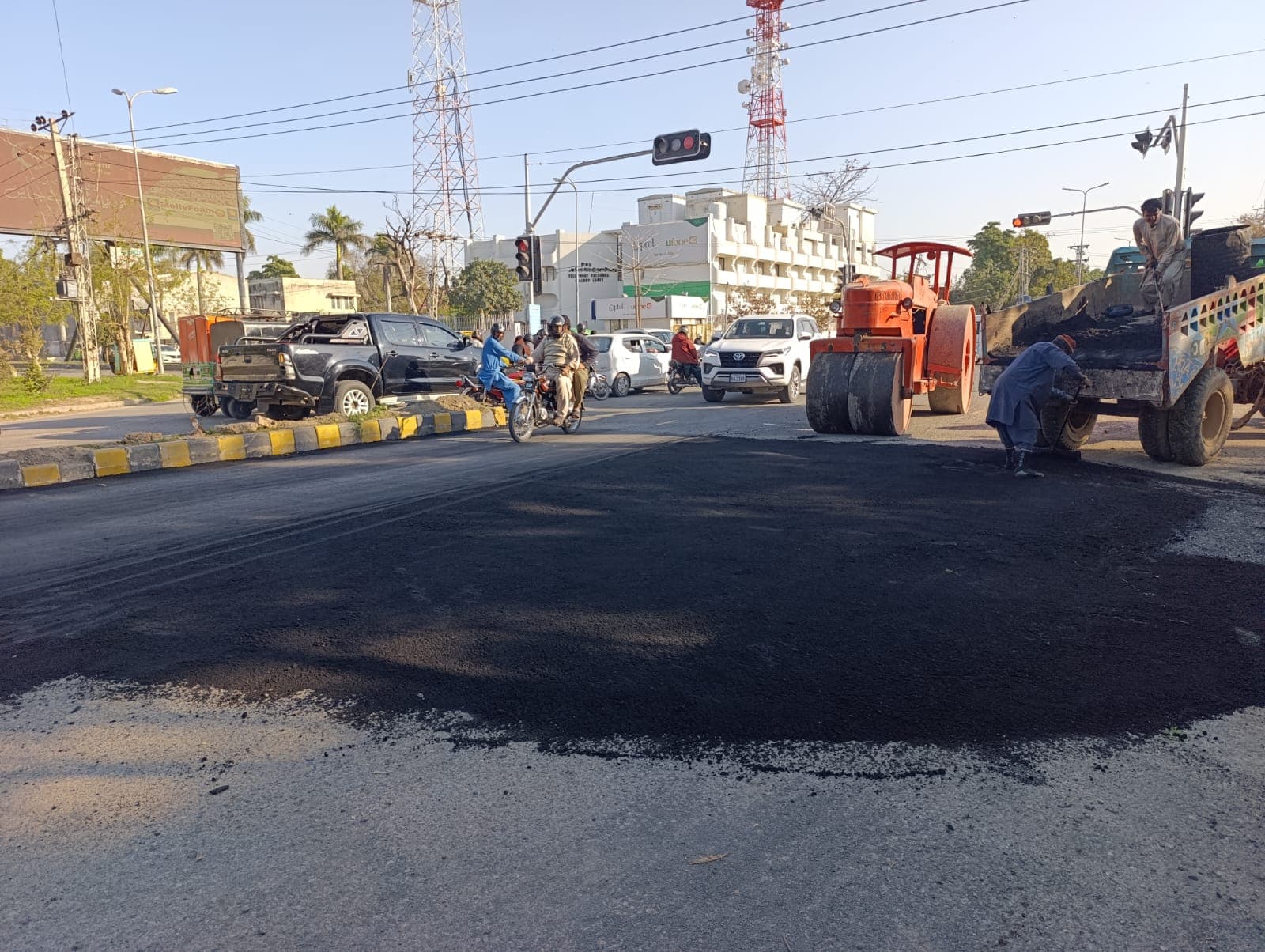 A rolled asphalt patch at a busy crossing, with the roller beside the traffic