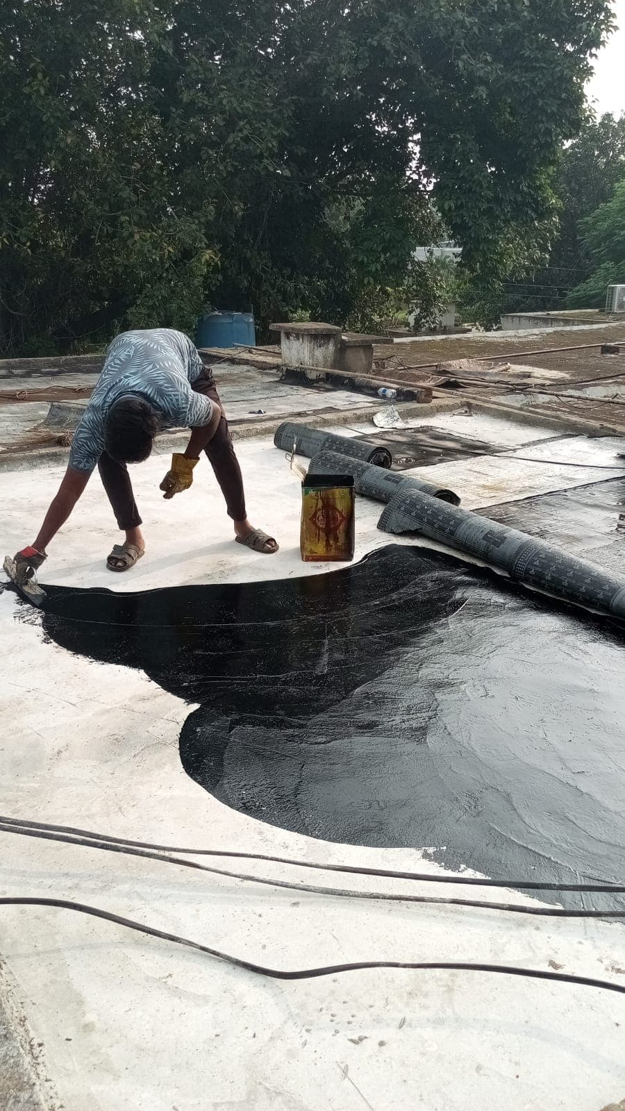 A worker brushing bitumen primer onto the roof beside rolls of membrane